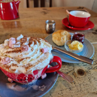 A large red cup on a blue saucer, full of hot chocolate topped with whipped cream and marshmallows. The hot chocolate has overflowed onto the saucer. Behind the drink is a scone on a plate with jam and butter, and behind that another red cup on a saucer next to a red tea pot. The table is an old wooden one.