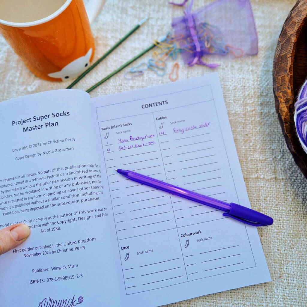 An open notebook with a purple pen lying across it is on a cream background next to an orange mug of tea, two green double pointed knitting needles, a small purple bag of bulb pins and a wooden dish with a ball of purple yarn in it