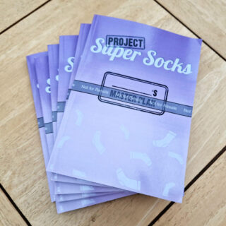 A pile of purple books with the words Project Super Socks printed on them is stacked on a wooden coffee table