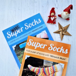 A blue book entitled More Super Socks and an orange book entitled Super Socks lying on a white board next to two Christmas ornaments and a brown paper star