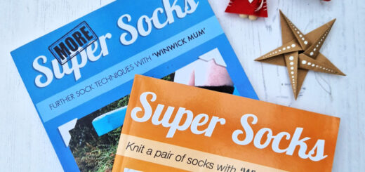 A blue book entitled More Super Socks and an orange book entitled Super Socks lying on a white board next to two Christmas ornaments and a brown paper star