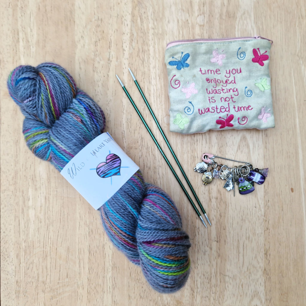 A skein of grey and rainbow yarn lies on a wooden coffee table next to two double pointed needles, some stitch markers and a small fabric notions pouch