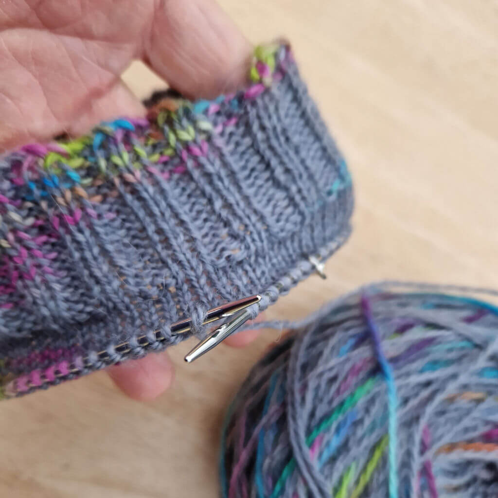 The cuff of a knitted sock showing how the mix of grey and rainbox colours has pooled to create a large area of grey