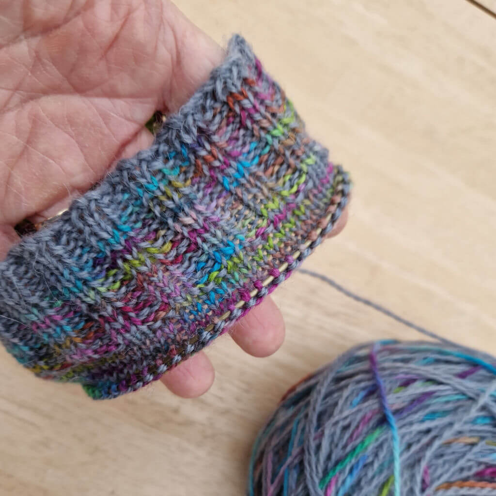 The cuff of a knitted sock showing how the mix of grey and rainbox colours has pooled to create a large area of grey