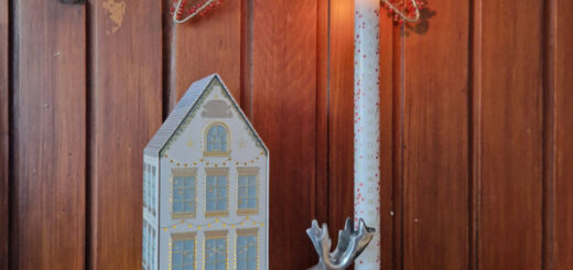 An Advent candle in a chrome reindeer-shaped holder next to a metal house-shaped light decoration. The back wall is wooden and there is a star hanging from it