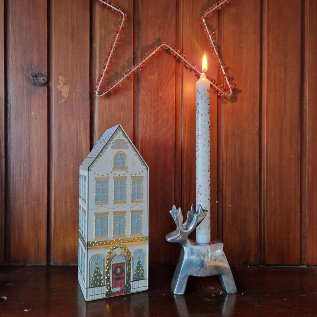 An Advent candle in a chrome reindeer-shaped holder next to a metal house-shaped light decoration. The back wall is wooden and there is a star hanging from it
