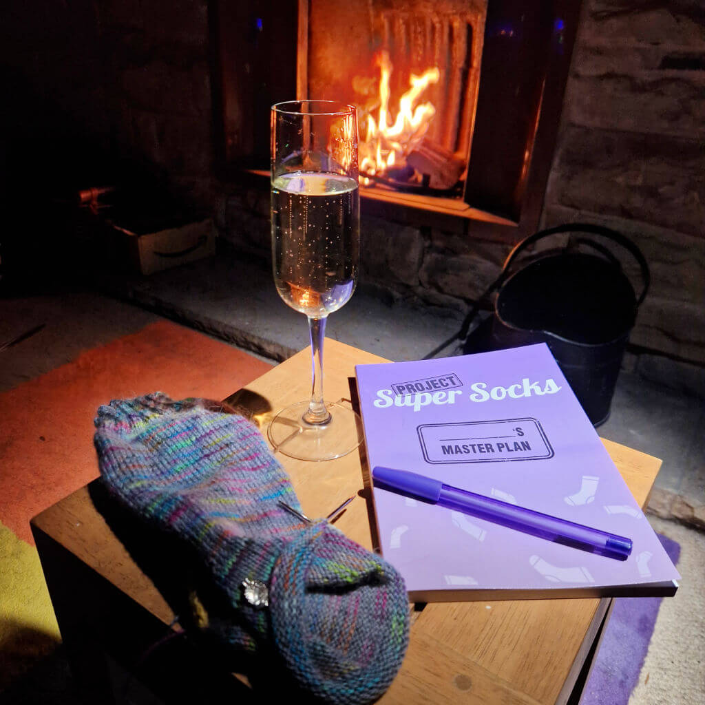 A purple book with a purple pen lying across it is on a wooden coffee table in front of an open fire. There is a glass of prosecco and a partly-knitted sock on the table too.