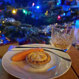 A small wooden coffee table with a white plate and a cut glass of whisky on it. There is a mince pie and a carrot on the plate and a Christmas tree decorated with lights in the background.