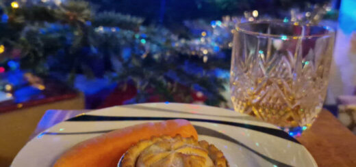 A small wooden coffee table with a white plate and a cut glass of whisky on it. There is a mince pie and a carrot on the plate and a Christmas tree decorated with lights in the background.