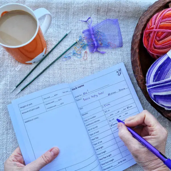 An open notebook with spaces to write information is lying on a fabric surface. Someone is writing in the book with a purple pen, and above it on the table is an orange mug of tea, two double pointed needles and a small bag of coloured bulb pins. There's a wooden bowl with ball of purple yarn and a ball of pink and red yarn in it to the right.