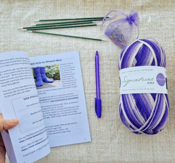 A book held open at a page showing a sock knitting pattern. Next to the book on a fabric background is a ball of purple striped yarn, a purple pen, five green double pointed needles and a small purple bag of coloured bulb pins