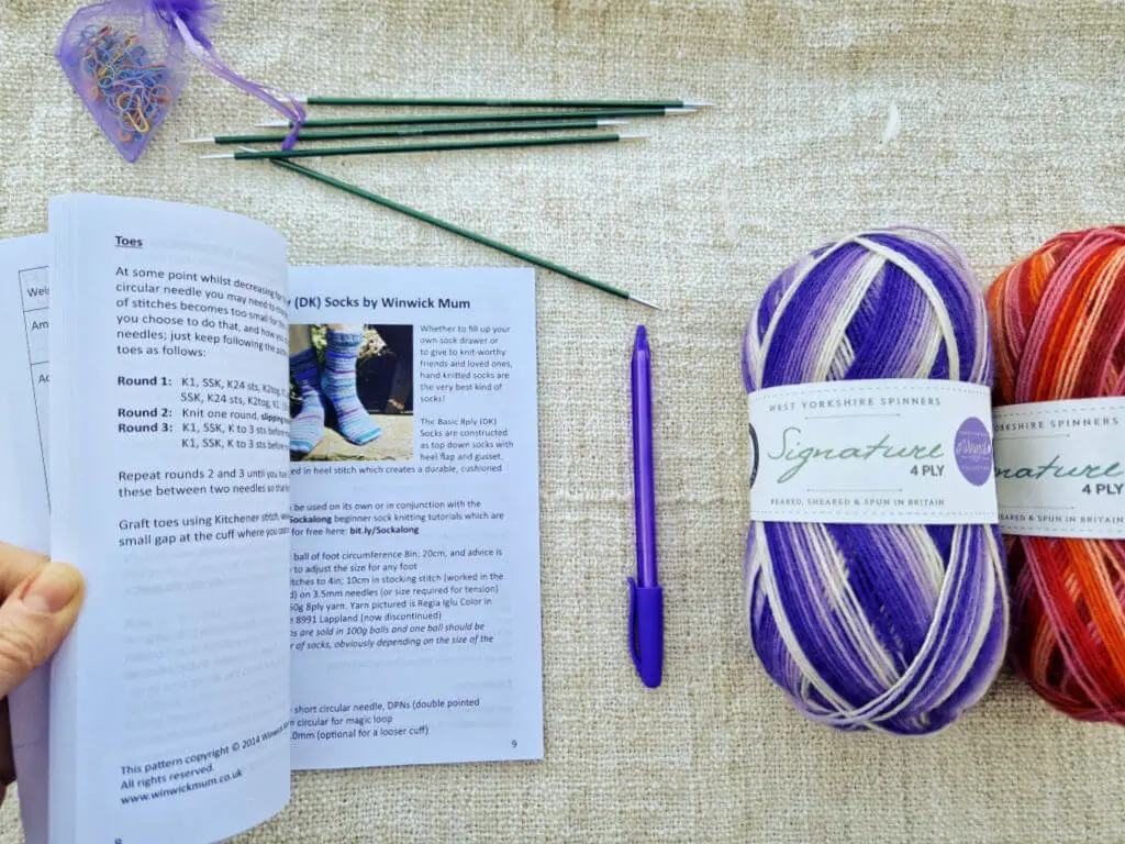 A book held open at a page showing a sock knitting pattern. Next to the book on a fabric background are two balls of striped yarn, one purple and one pink and red, a purple pen, five green double pointed needles and a small purple bag of coloured bulb pins