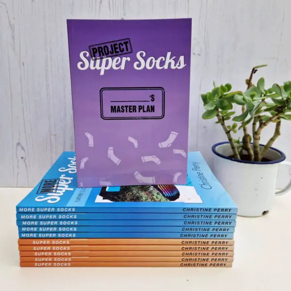 A purple book with the title "Project Super Socks" is balanced on a stack of blue and orange books against a wooden background. There's a pot plant in the right hand corner