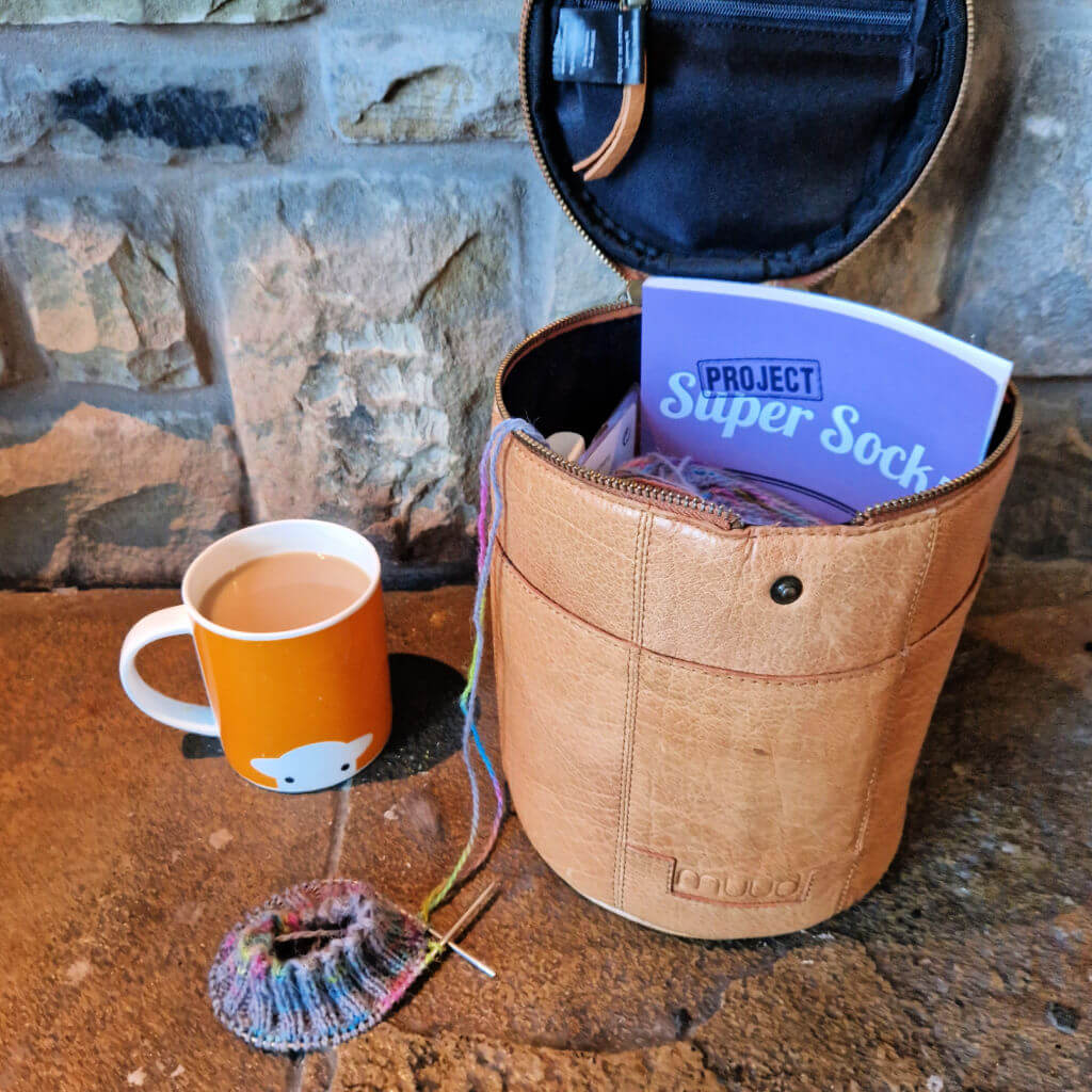 A round leather knitting bag sits on a stone hearth next to an orange mug of tea and a partly knitted sock. There is a purple book in the knitting bag and it fits perfectly.