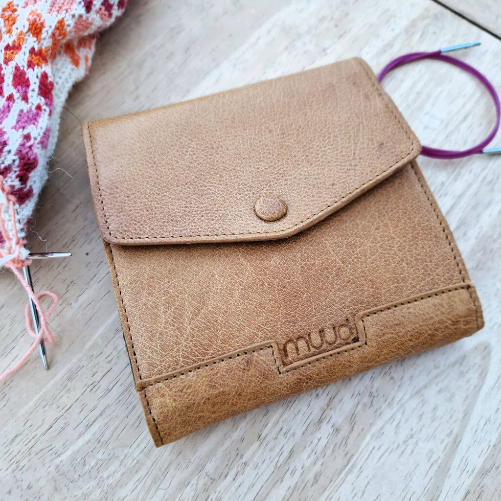 A square brown leather case which folds over itself to close with a press stud is lying on a wooden coffee table next to a circular needle cable and a partly knitted colourwork sock in s pink, red and cream stripes
