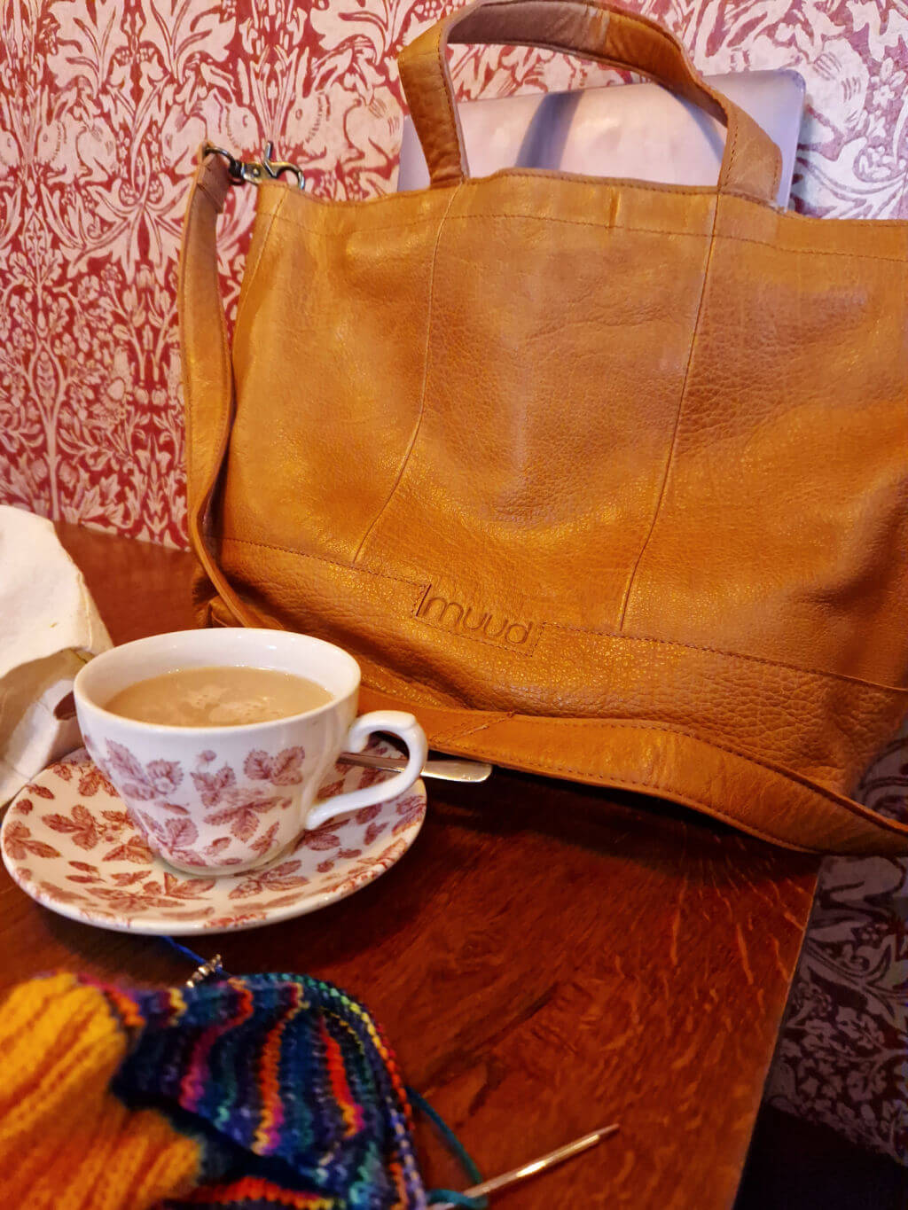 A brown leather shopper bag on a table next to a cup of tea in a china cup and saucer, and a partly knitted sock. The pattern on the teacup is similar to the pattern on the wallpaper behind the bag