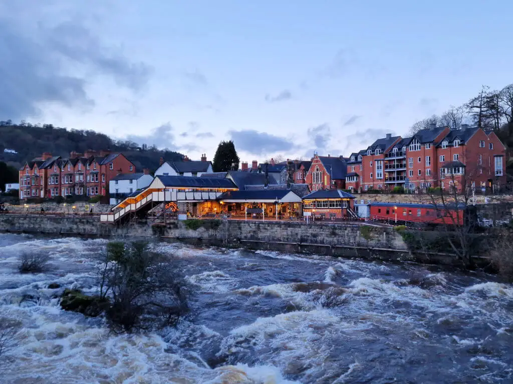 A view across a raging river to where a railway station is lit up as the daylight fades