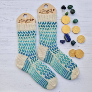 A pair of blue striped and cream colourwork socks on wooden sock blockers lying on a white wooden board. There are gold chocolate coins and gemstones in shades of blue and green lying next to the socks.
