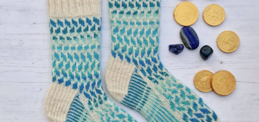 A pair of blue striped and cream colourwork socks on wooden sock blockers lying on a white wooden board. There are gold chocolate coins and gemstones in shades of blue and green lying next to the socks.