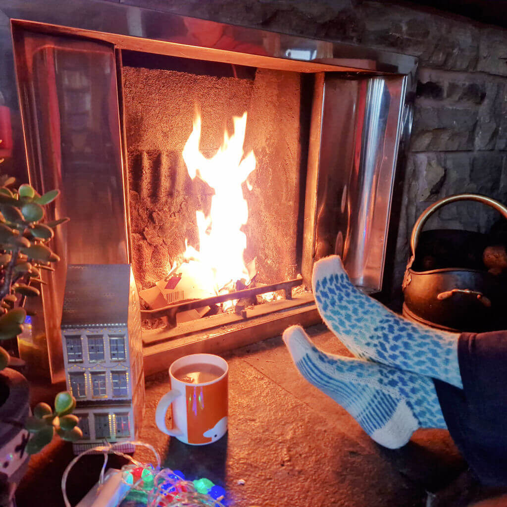 A pair of blue and cream colourwork socks modelled on feet resting on a stone hearth in front of an open fire