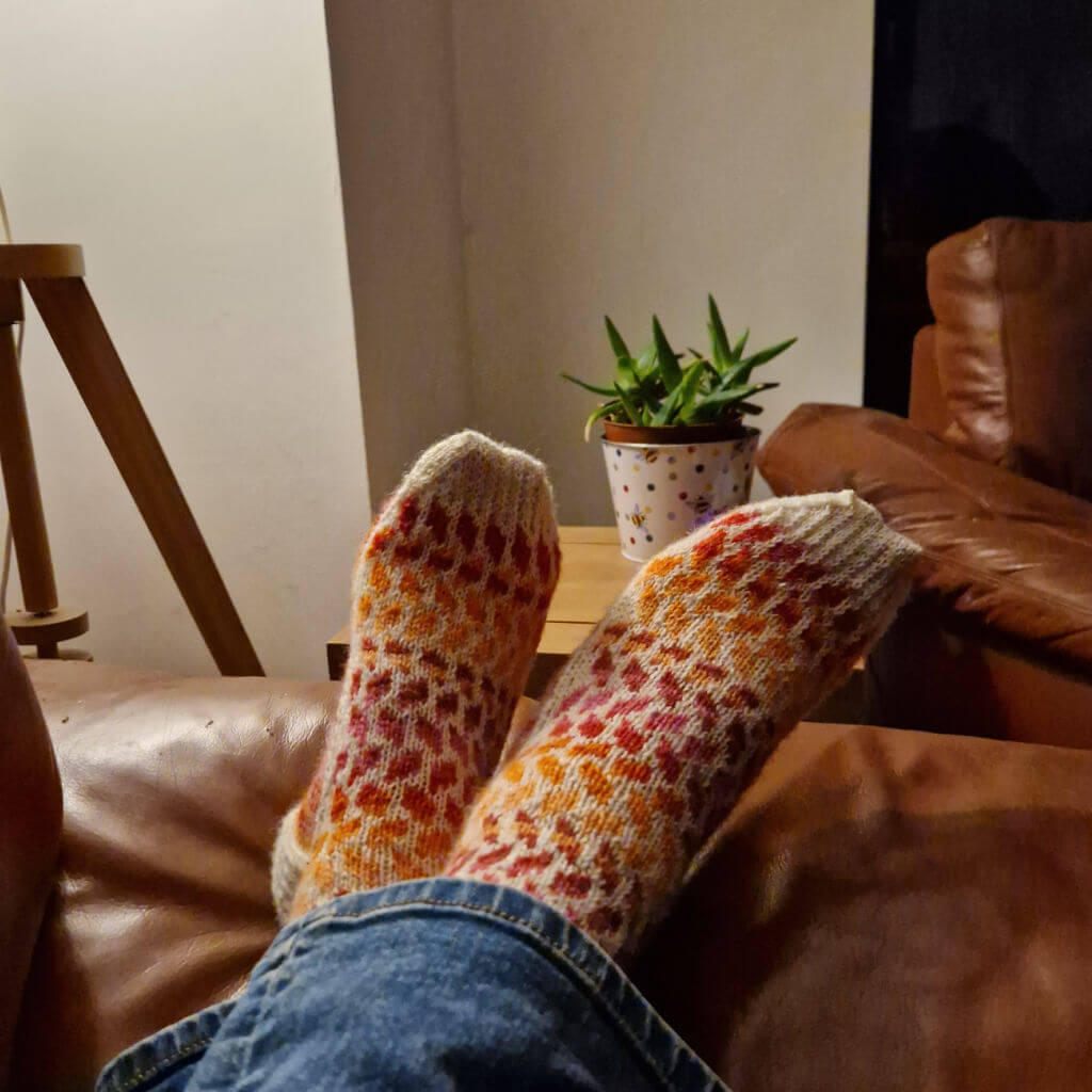 A pair of cream, pink, orange and red striped colourwork socks modelled on feet. The feet are resting on the arm of a sofa with an armchair and coffee table with a plant on it in the background
