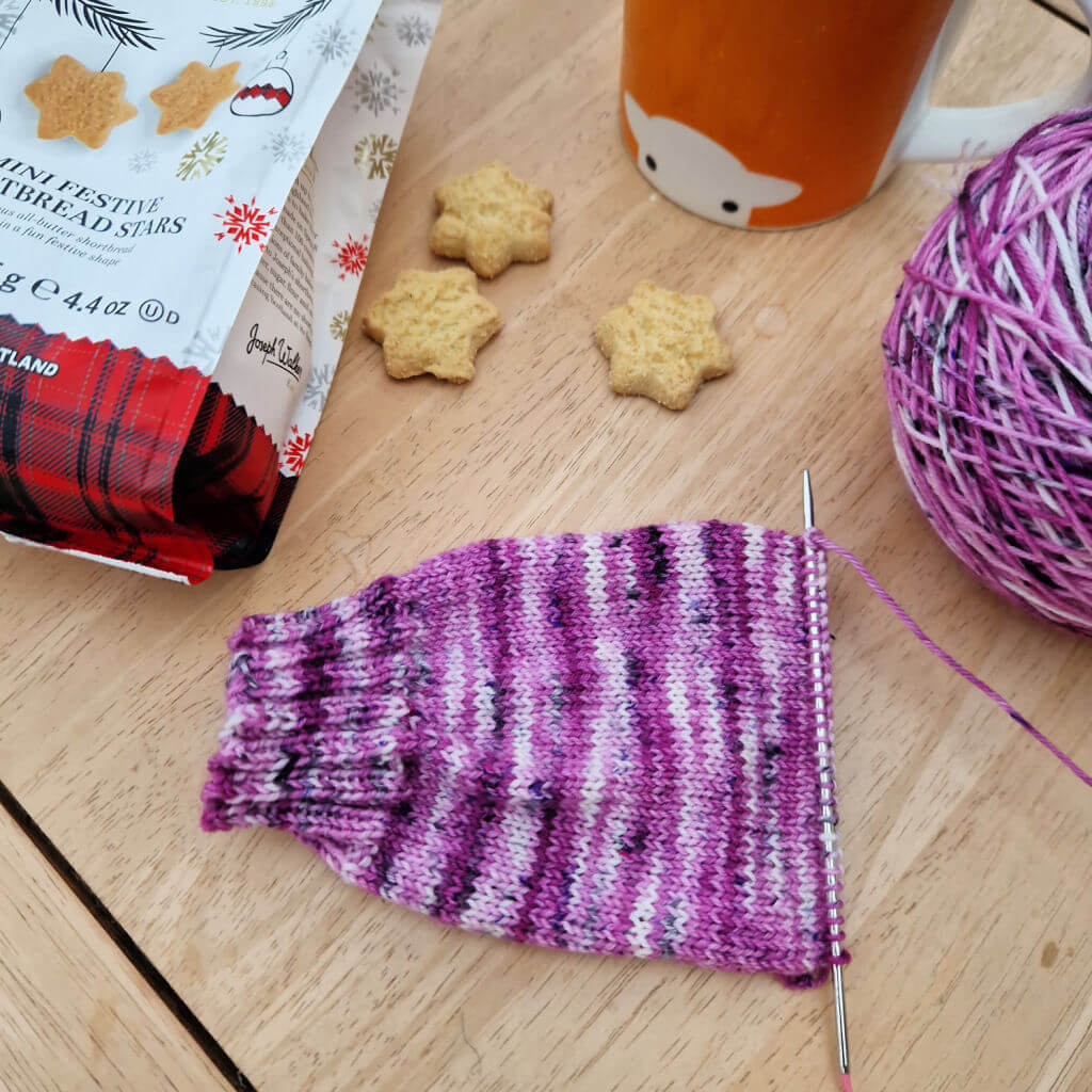 A partly-knitted sock in purple variegated yarn on a wooden coffee table next to the ball of yarn, an orange mug of tea and an open packet of shortbread stars