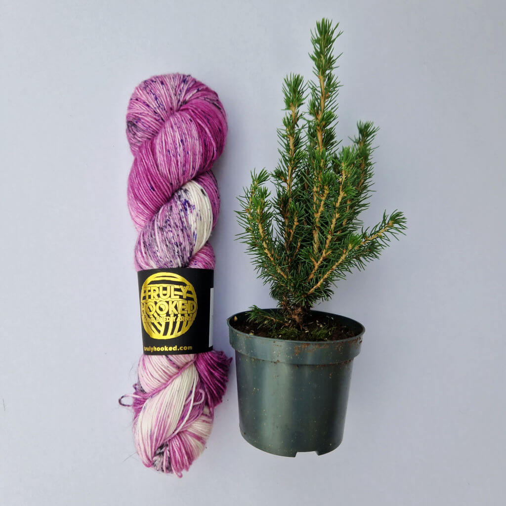 A skein of pink speckled yarn and a small fir tree in a pot are lying next to each other on a grey background