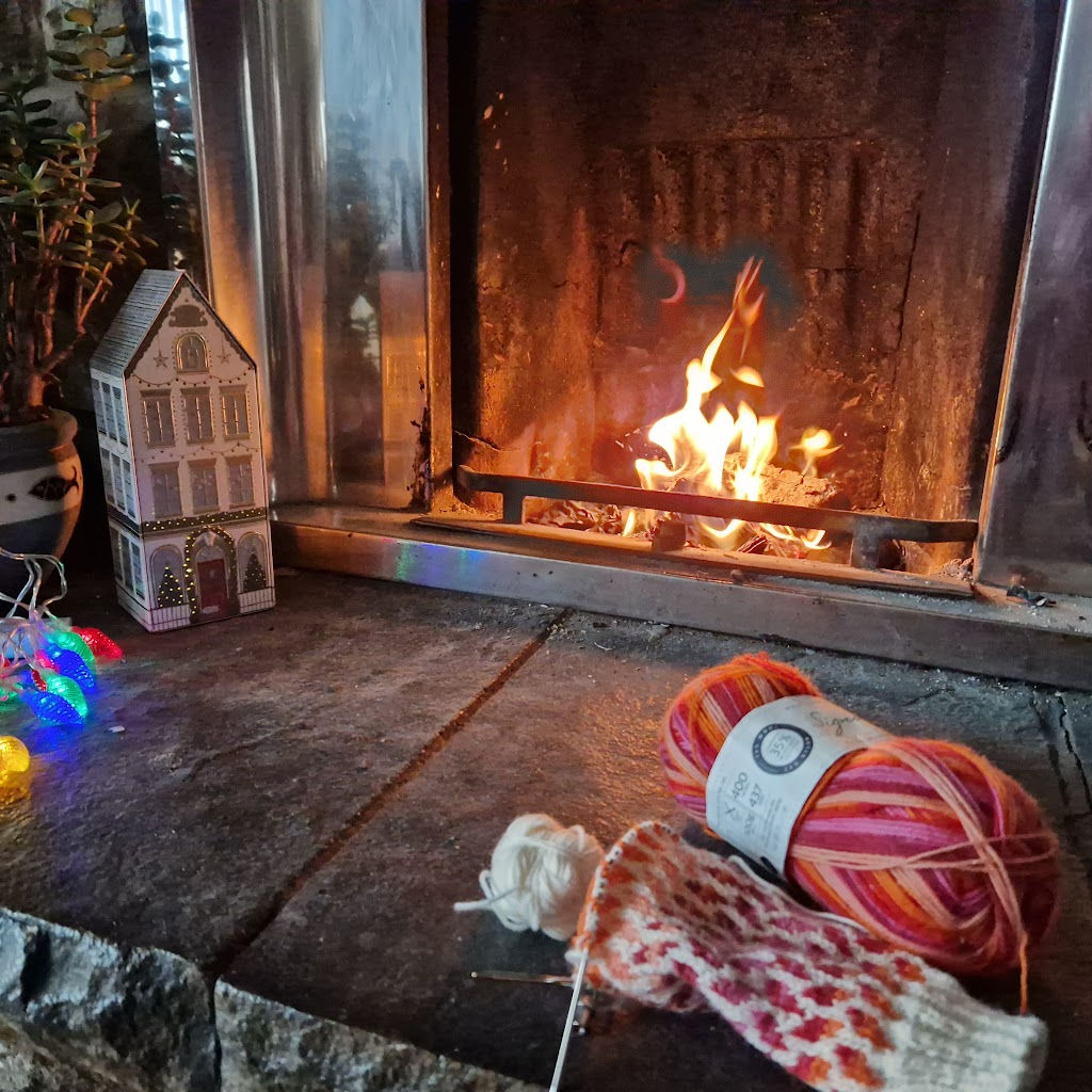 A partly knitted pink, red and cream colourwork sock on a stone hearth next to an open fire