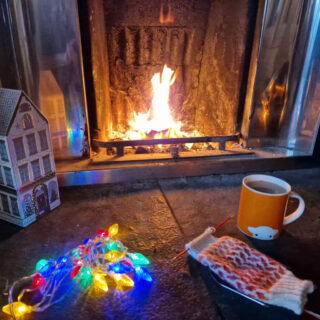 A partly knitted pink, red and cream colourwork sock on a stone hearth next to an open fire