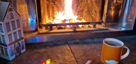 A partly knitted pink, red and cream colourwork sock on a stone hearth next to an open fire
