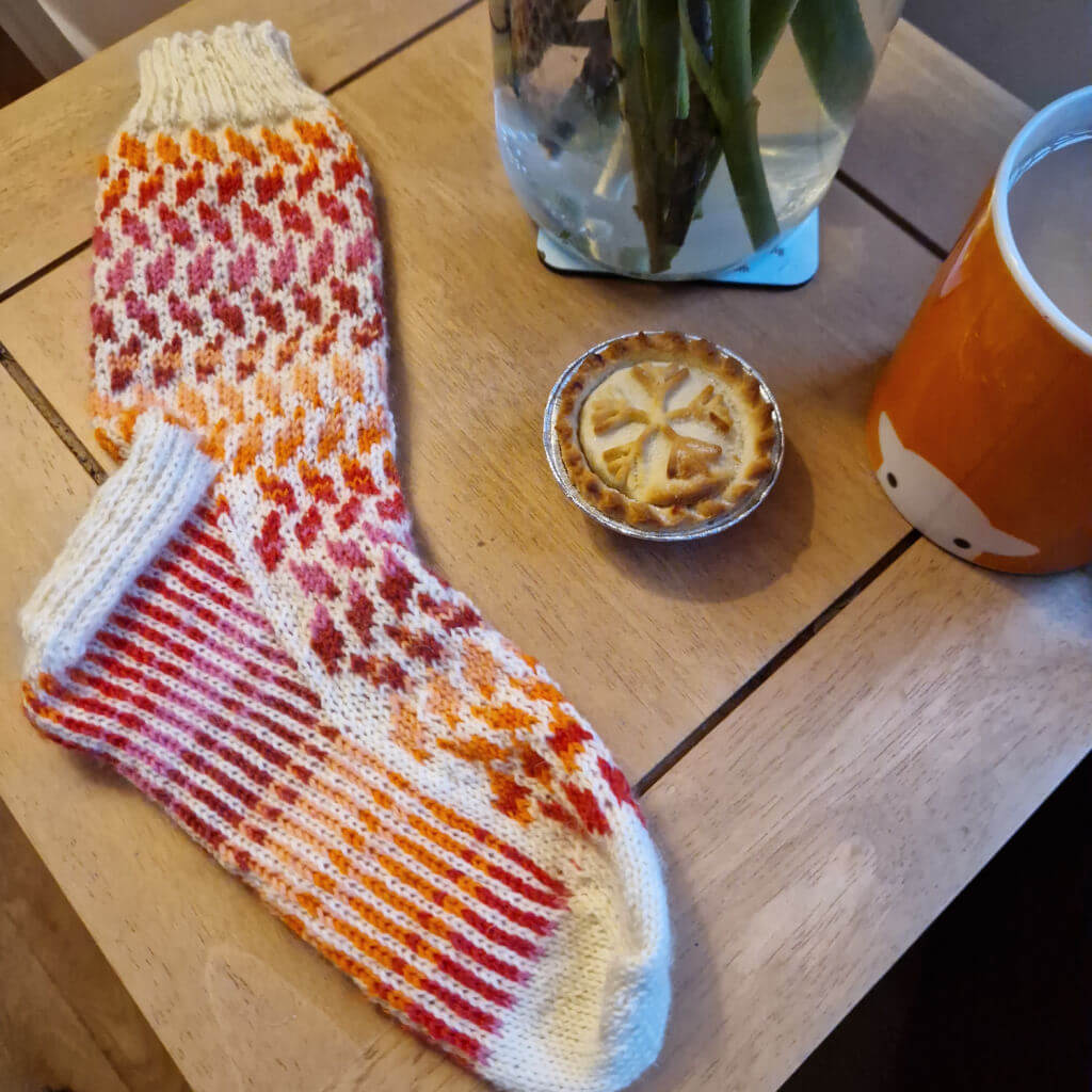 A pink and cream striped colourwork sock is lying on a coffee table next to a mince pie, an orange mug of tea and a vase of flowers