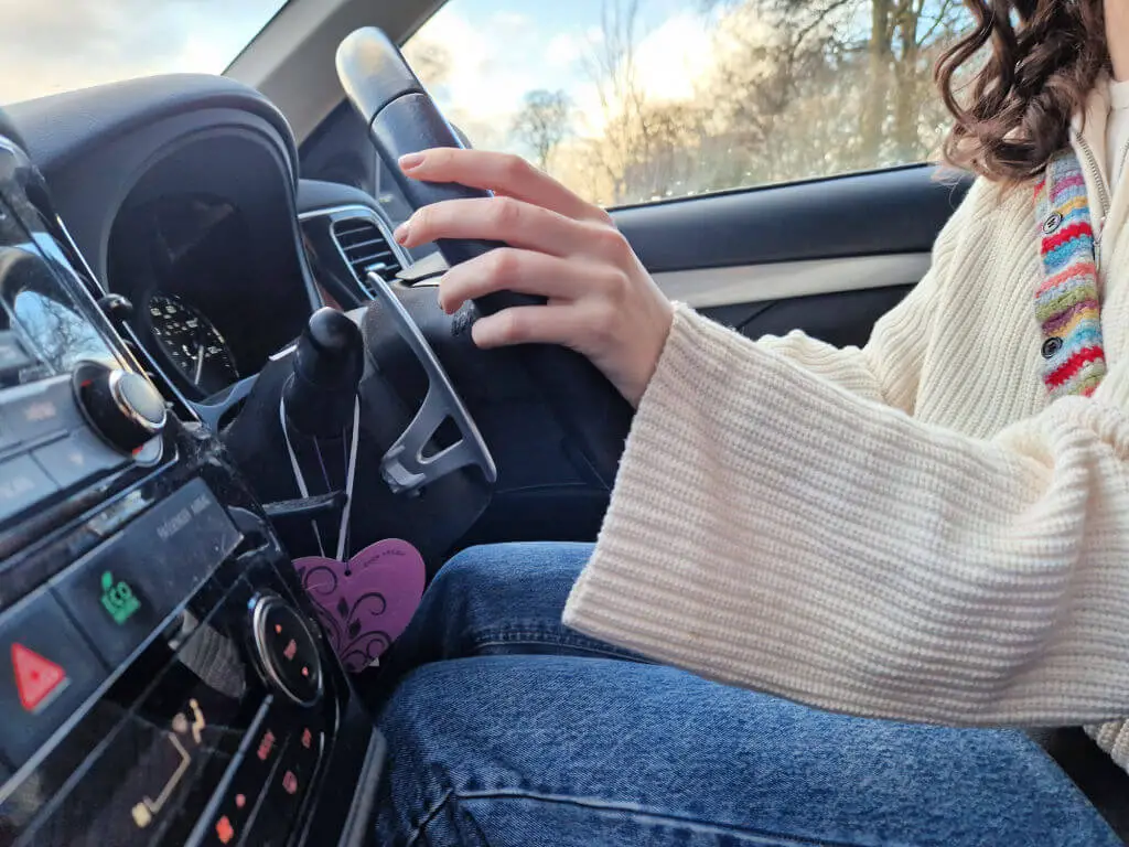A young woman with brown hair and wearing a white jumper is driving a car