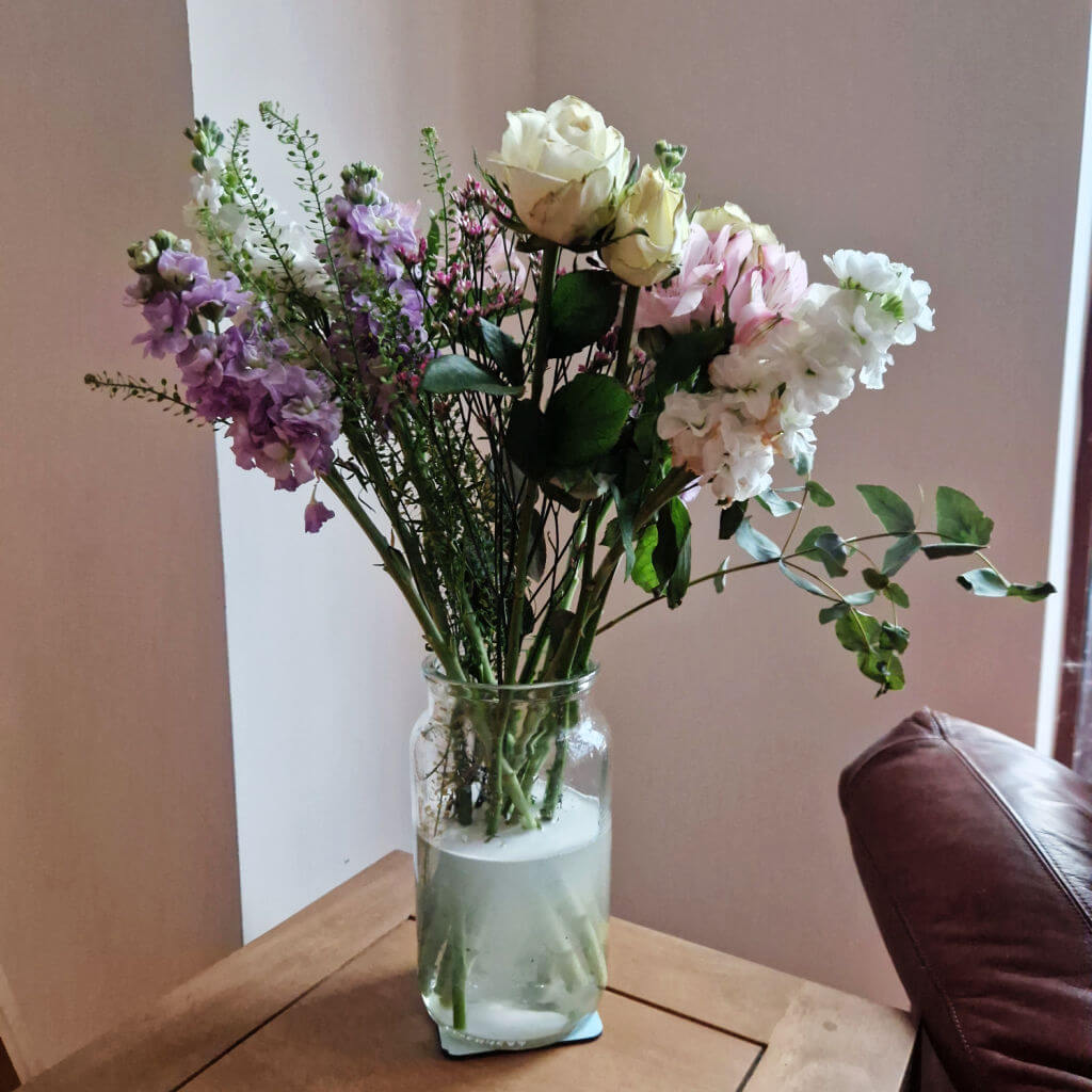 A glass vase of flowers in pastel shades of white, pink and purple on a wooden coffee table in a corner, next to a brown leather armchair