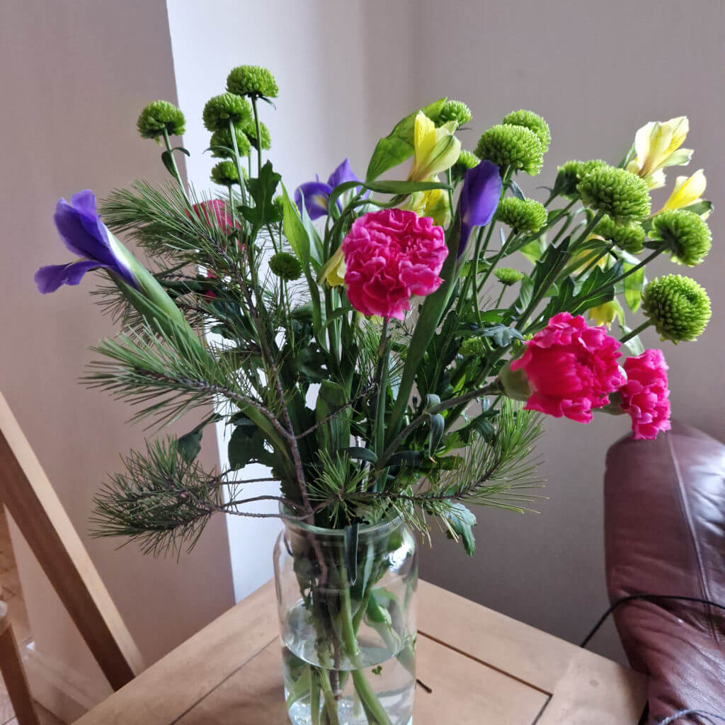 A vase of cut flowers in shades of pink, blue and yellow with green leafy fronds as well