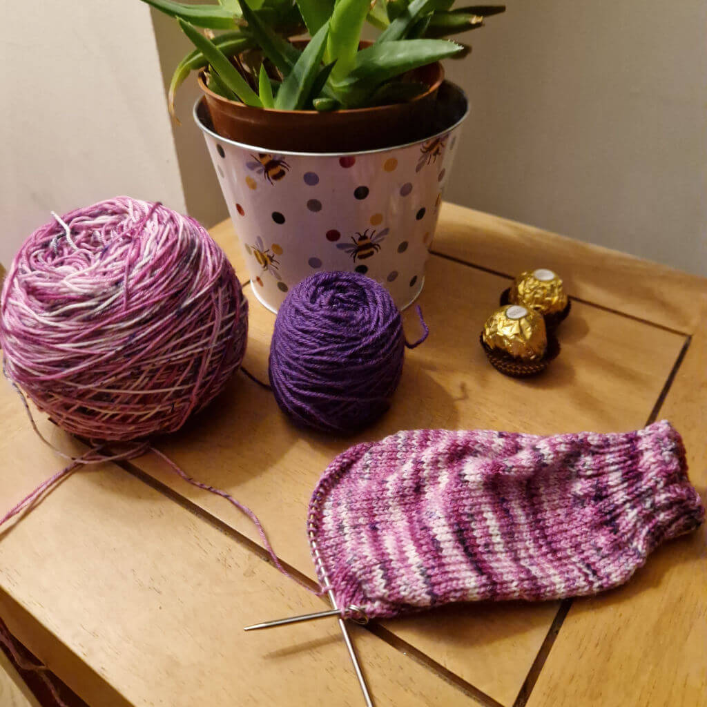 A partly-knitted pink variegated sock resting on a wooden coffee table next to a potted plant in a metal planter decorated with spots and bees. There are two round chocolates in gold wrappers on the table as well.