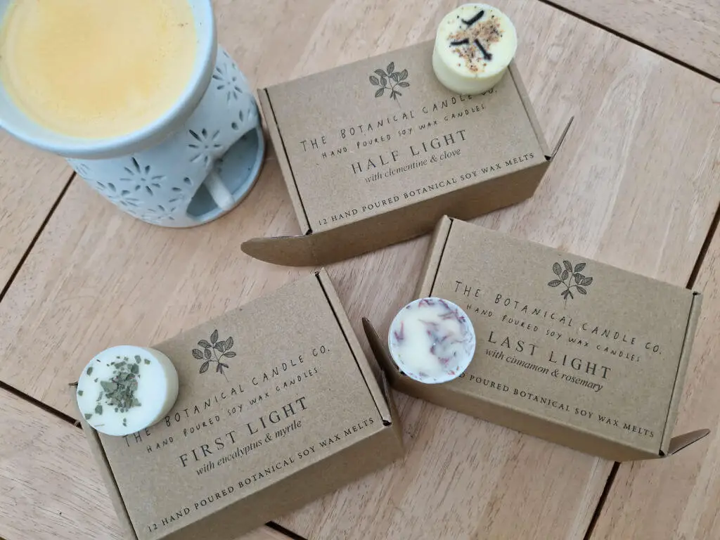 Three small cardboard boxes containing wax melts on a wooden coffee table next to a wax melt burner. There is a round wax melt from each box on the box it came out of