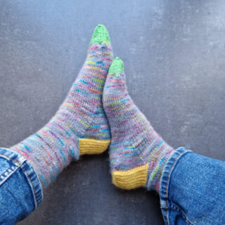 A pair of hand knitted socks in grey and rainbow yarn with yellow heels and green toes, modelled on feet against a tiled floor