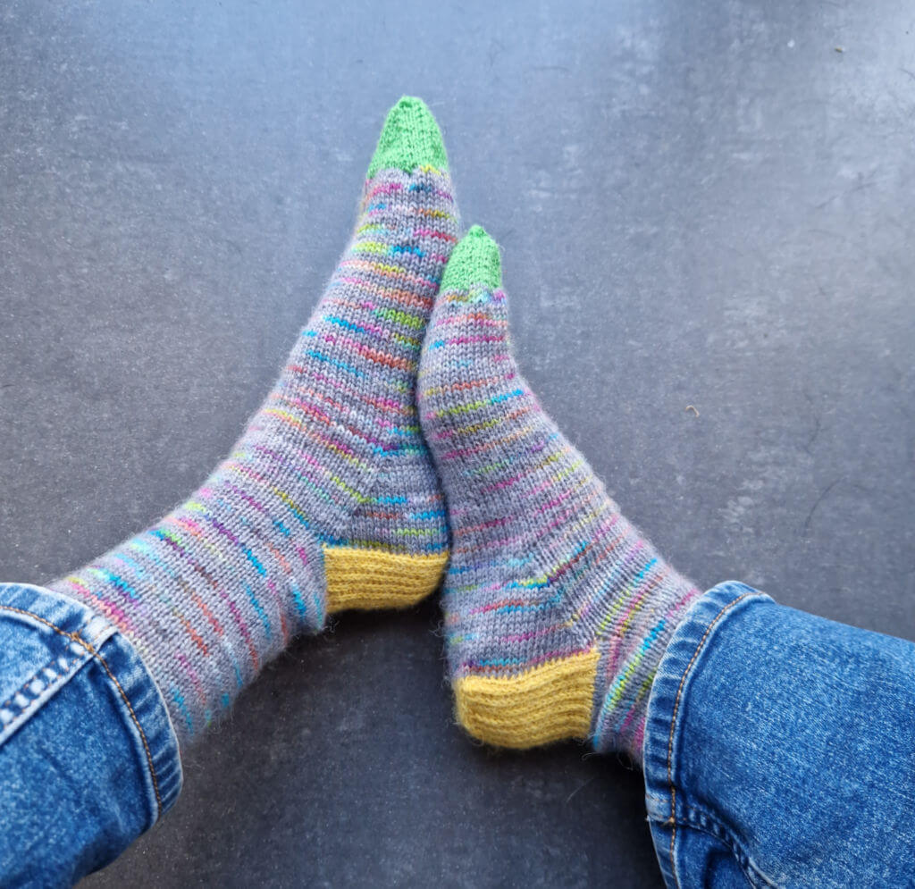 A pair of hand knitted socks in grey and rainbow yarn with yellow heels and green toes, modelled on feet against a tiled floor