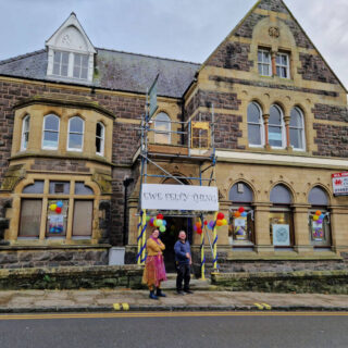 A man and a woman standing outside an old brick building with ornate windows. The sign above their heads, attached to scaffolding, reads "Ewe Felty Thing"