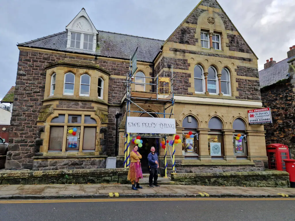 A man and a woman standing outside an old brick building with ornate windows. The sign above their heads, attached to scaffolding, reads "Ewe Felty Thing"