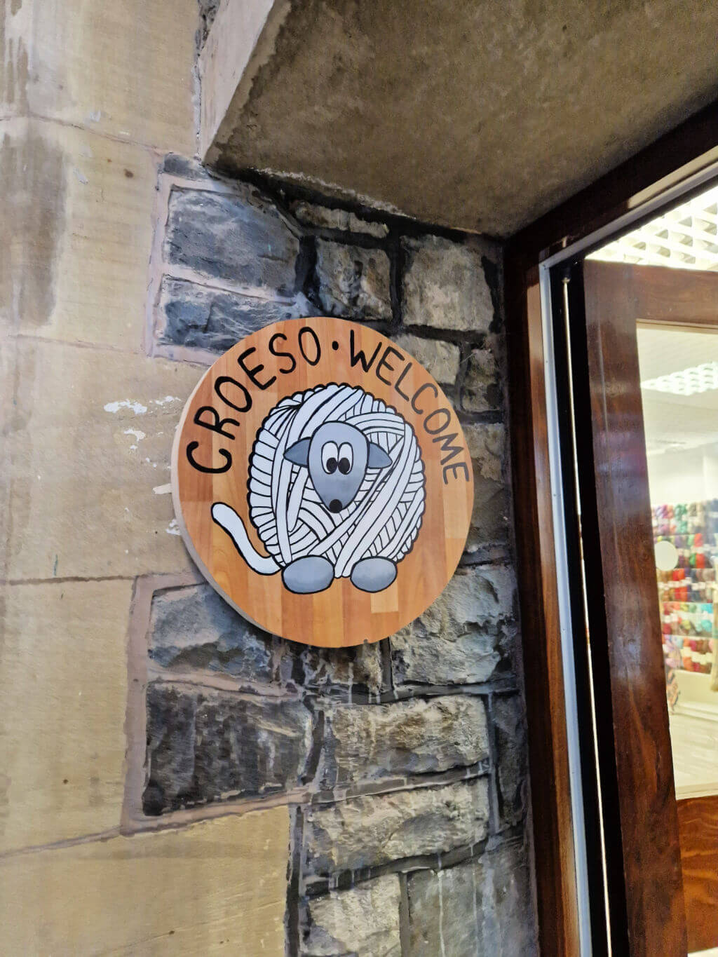 A round wooden sign fixed to the stone wall of a shop entrance. There is a drawing of a sheep made from a ball of yarn and the words Croeso - Welcome on the sign