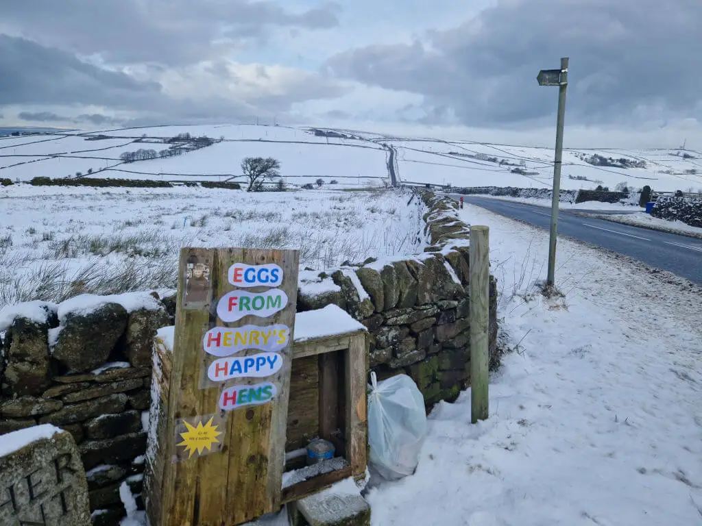 A wooden honesty box for eggs. The words "Eggs from Henry's Happy Hens" are on the box. Beyond is a view across snowy moorland 