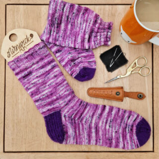 A pair of pink socks with deep purple heels and toes lie on a wooden coffee table next to a leather scissors pouch, a pair of gold bird-shaped scissors, two wool needles on a piece of a black fabric and an orange mug of tea