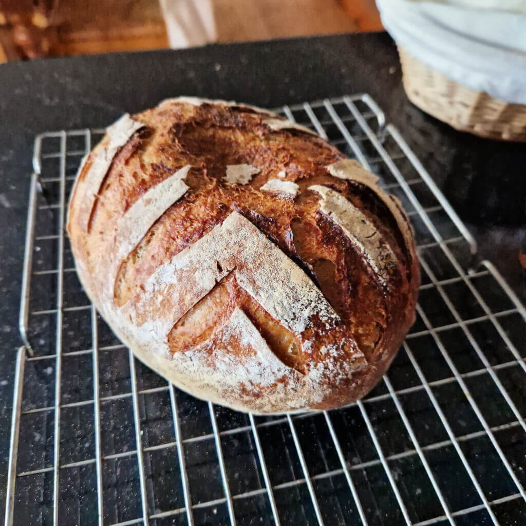 A sourdough loaf with a pattern cut into the crust sits on a wire baking rack on a kitchen worktop