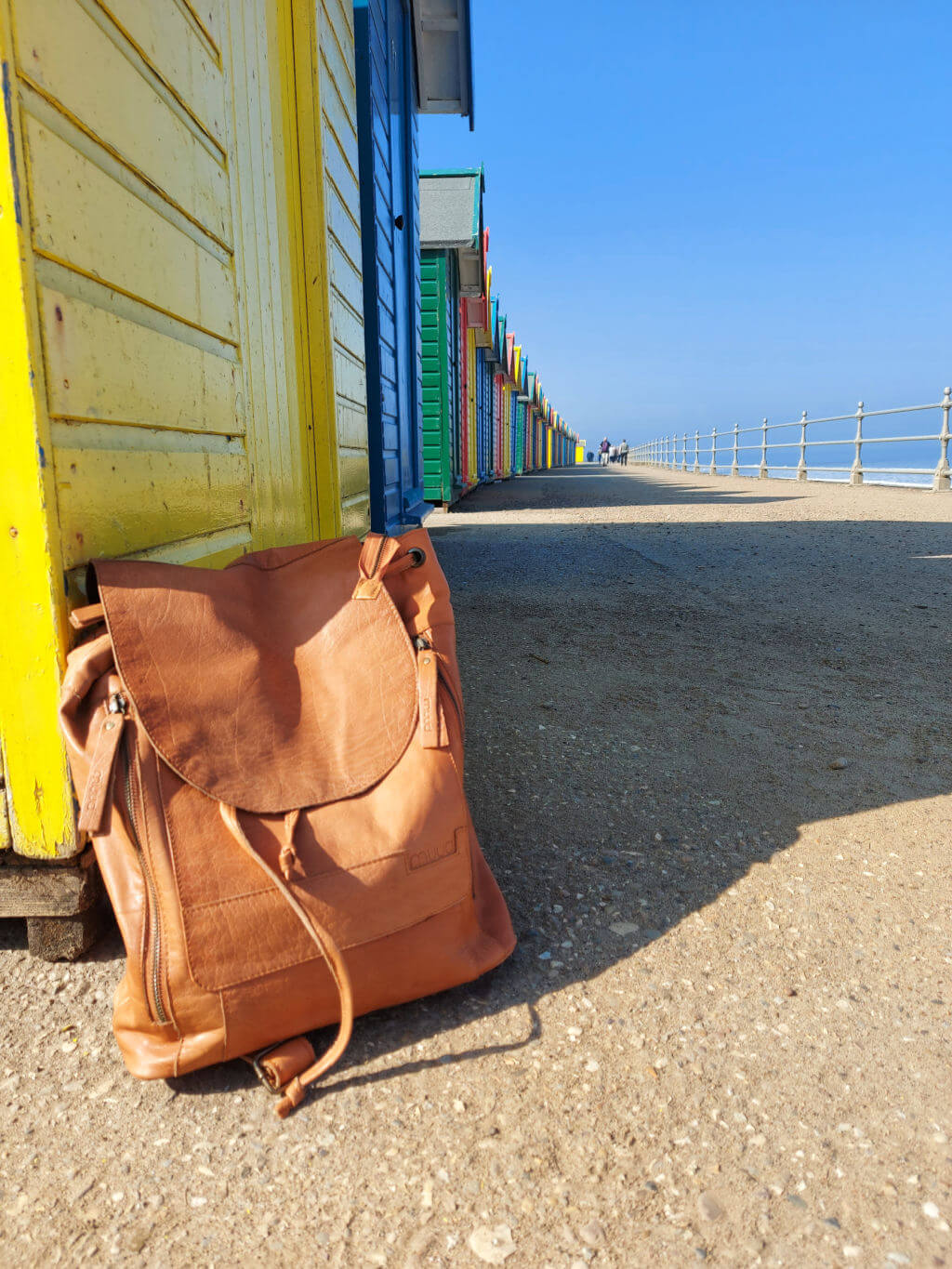 A brown leather Muud Gimo backpack bag leaning on the yellow wall of a beach hut. The line of coloured beach huts stretches out into the distance