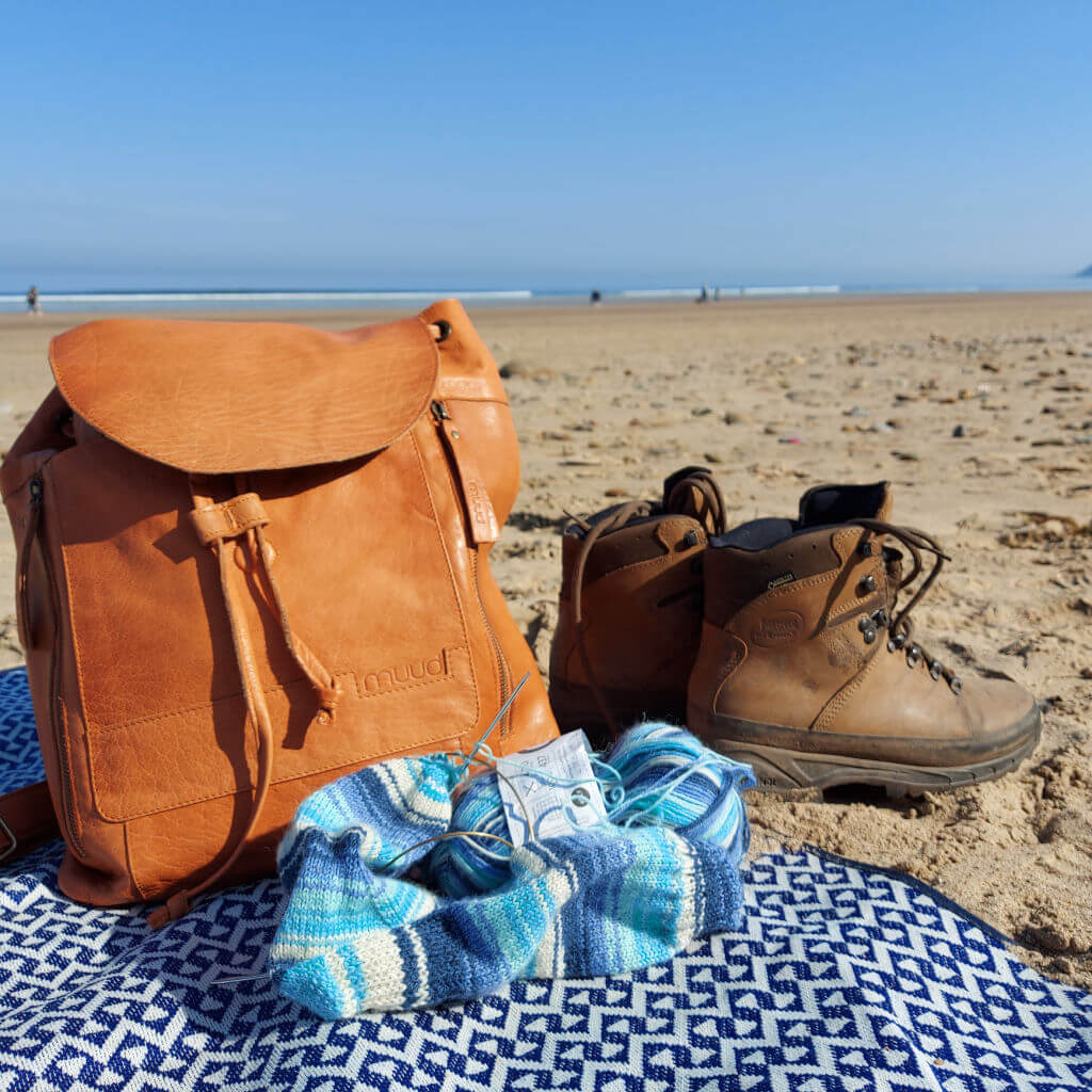 A brown leather Muud Gimo backpack on a patterned beach blanket next to a pair of partly-knitted blue striped socks and a pair of walking boots. The boots are on the sand and behind is the view to the sea