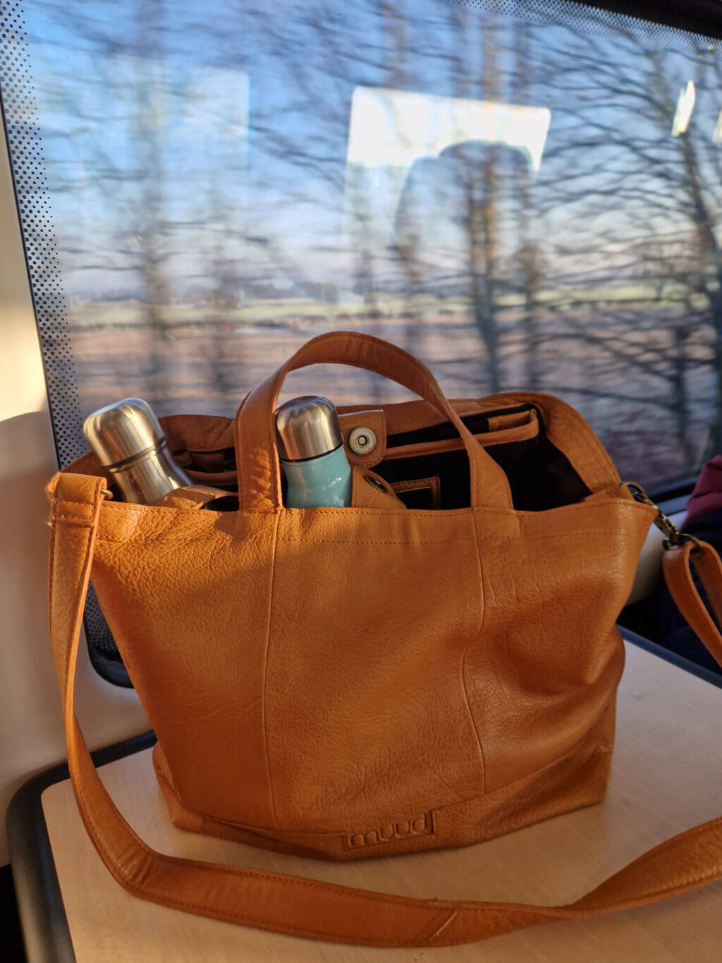 A brown leatehr Muud Hiba bag on a train table. There are two aluminium water bottles in the bag and the scenery is rushing past the window behind