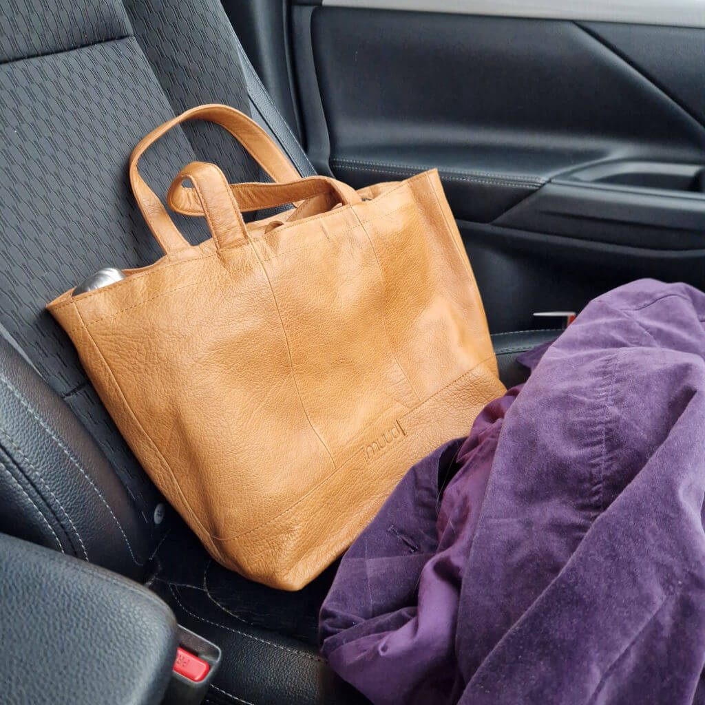 A brown leather Muud Hiba bag on a car seat next to a purple coat