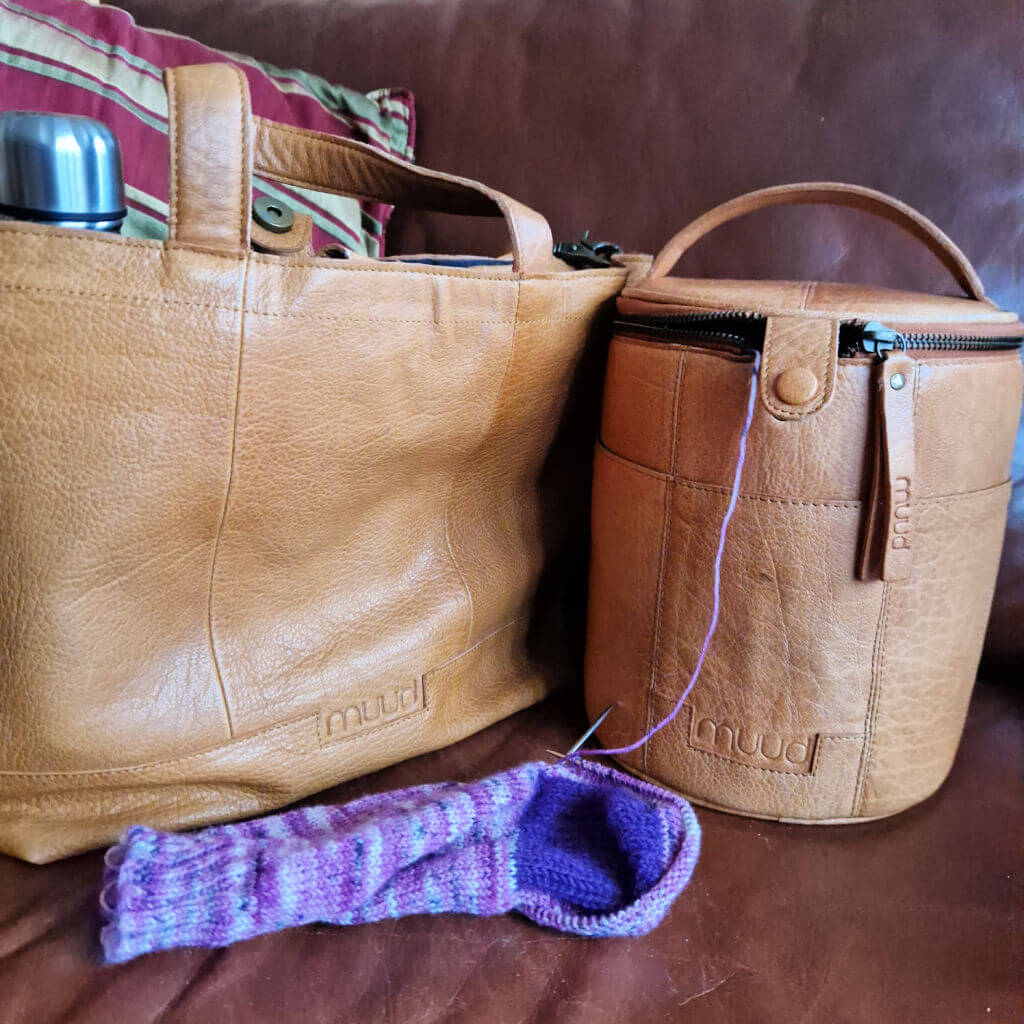 A brown leather Muud Hiba bag on a brown leather sofa next to a brown leather Muud Saturn knitting bag and a partly-knitted purple sock