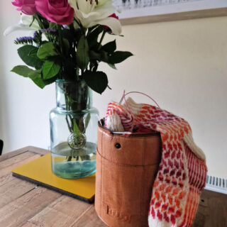 A partly-knitted cream, pink, red and orange colourwork sock resting on a brown leather Muud Saturn knitting bag sits on a table next to a vase of flowers. The picture on the wall behind has the word "beautiful" on it
